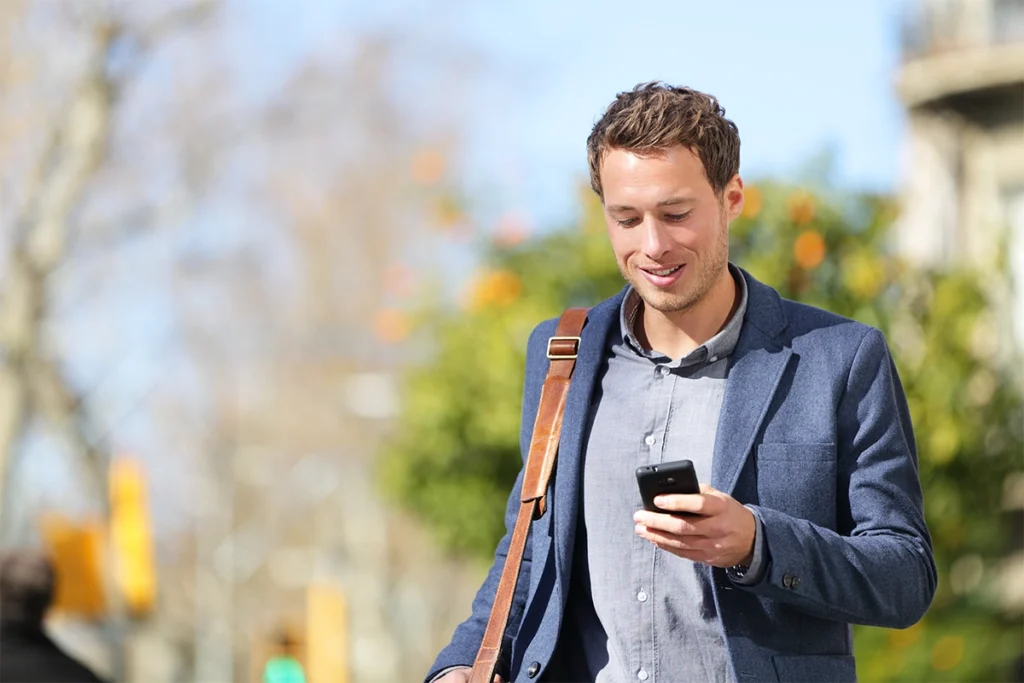 junger Mann mit dem Blick aufs Smartphone und einer Ledertasche über der Schulter.