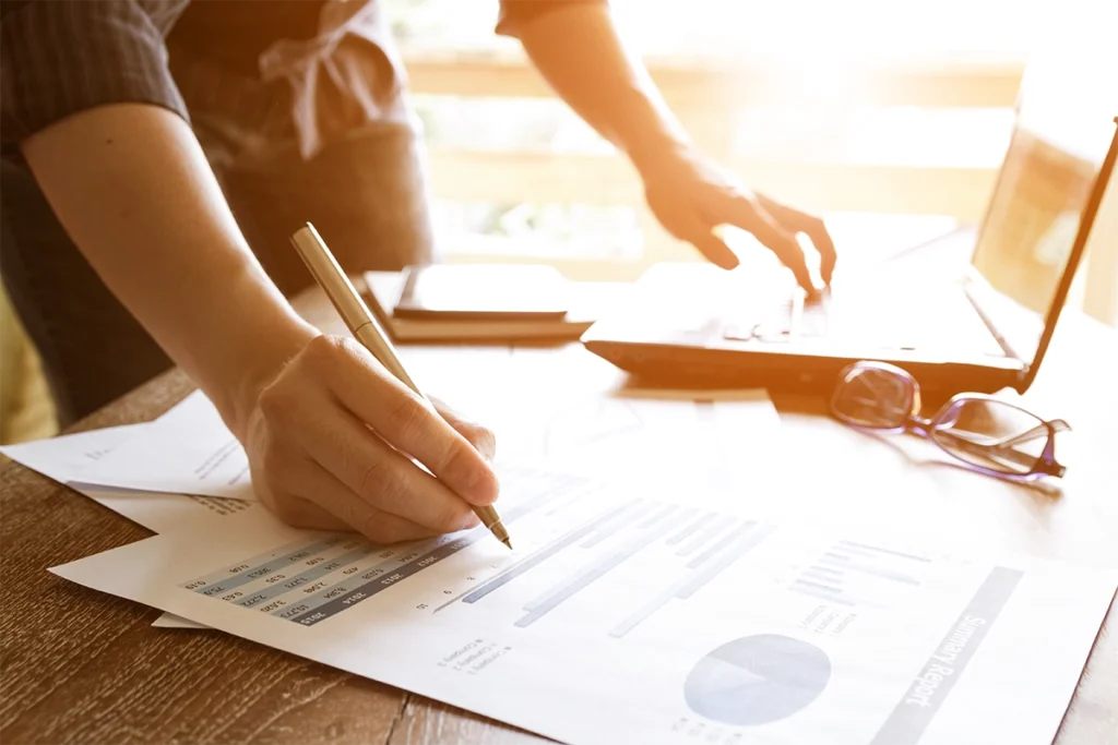 Eine Person mit einer Hand am Laptop und einem Stift um etwas abzugleichen. Neben dem Laptop liegt eine Brille.